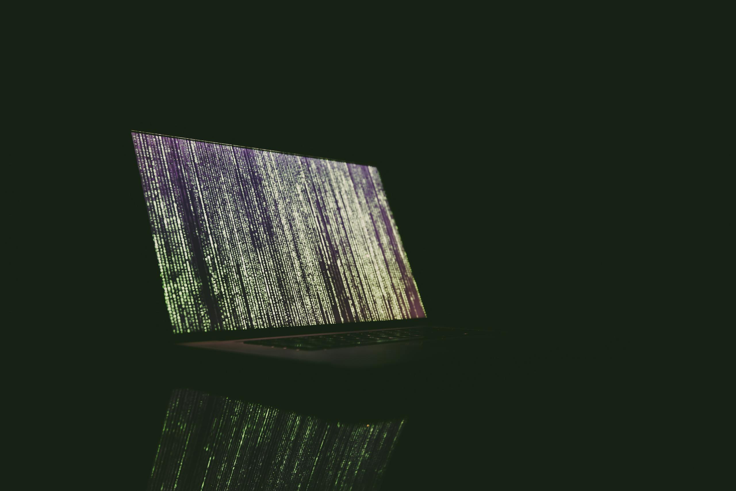 Whatsontech A laptop displaying green and purple code in a dark room, reflecting on a surface.