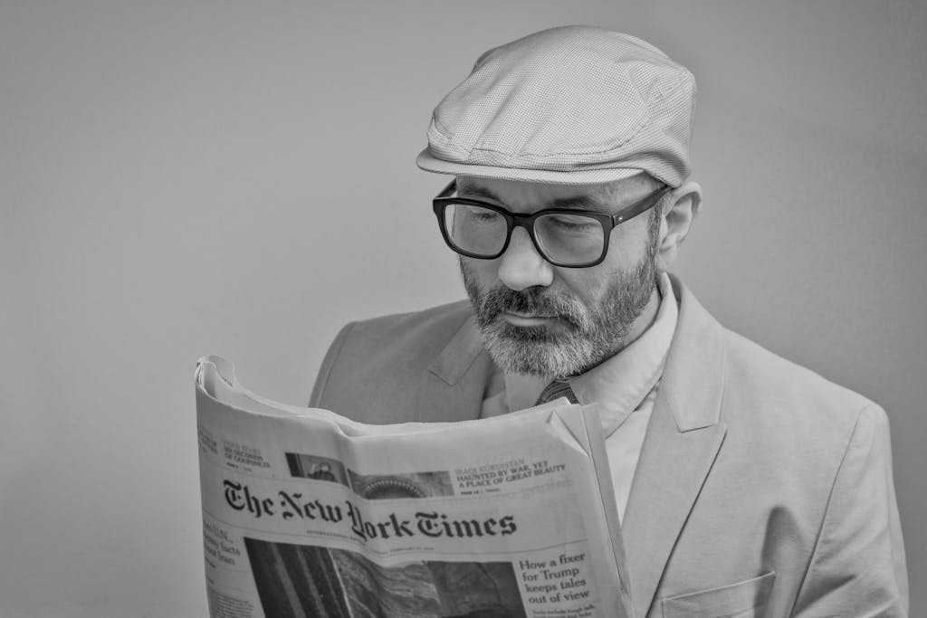 Newstook A man in a suit, wearing glasses and a cap, reads The New York Times.