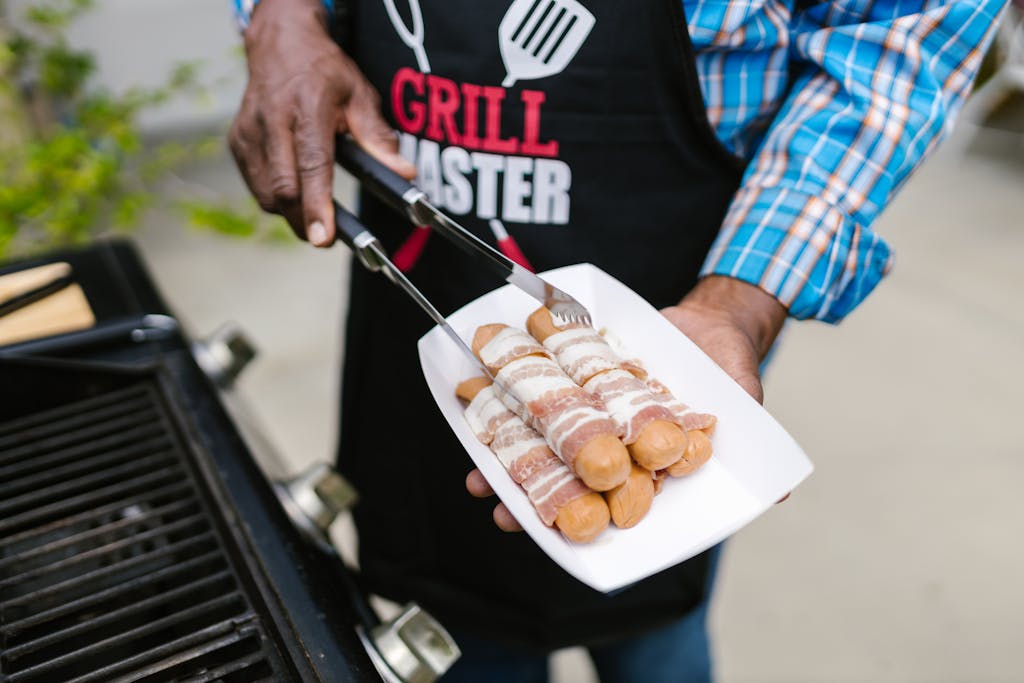 Weber Grillvorführung A person in an apron grilling bacon-wrapped hotdogs outdoors on a sunny day.
