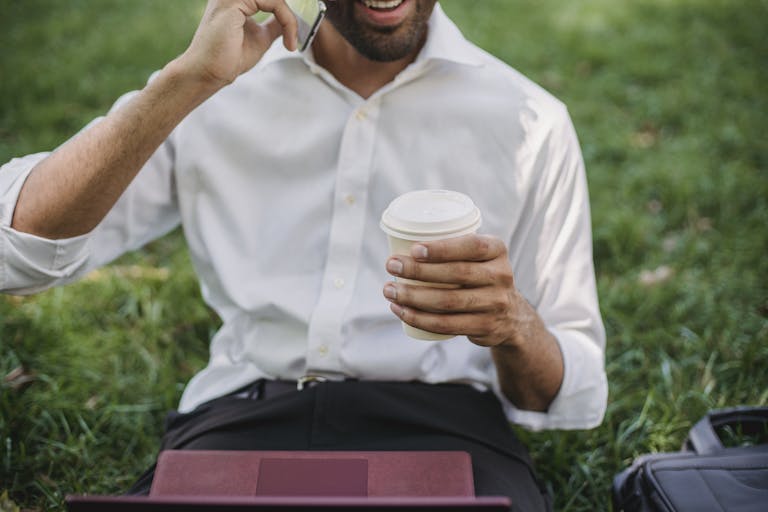 Entrepreneur Break A smiling man in a white shirt holding coffee and talking on the phone outdoors.