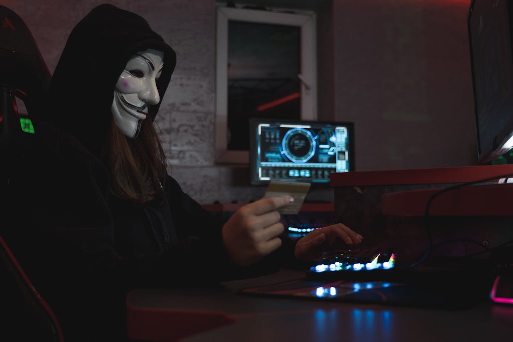 Whatsontech Masked hacker with credit card at computer, symbolizing cybercrime and anonymity.