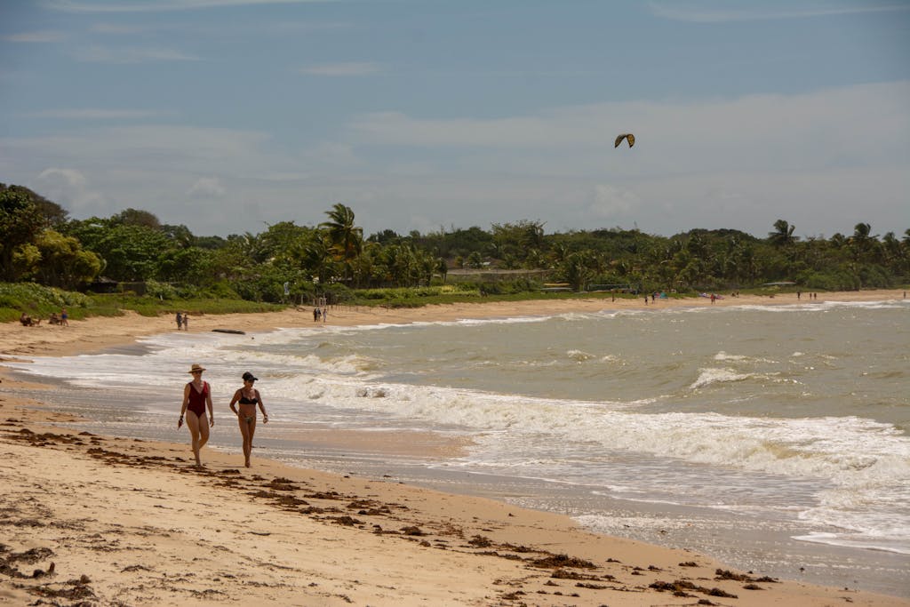 Juntos Seguros I Two women walking on a sunny beach in Porto Seguro, Brazil, vibrant tropical scenery.