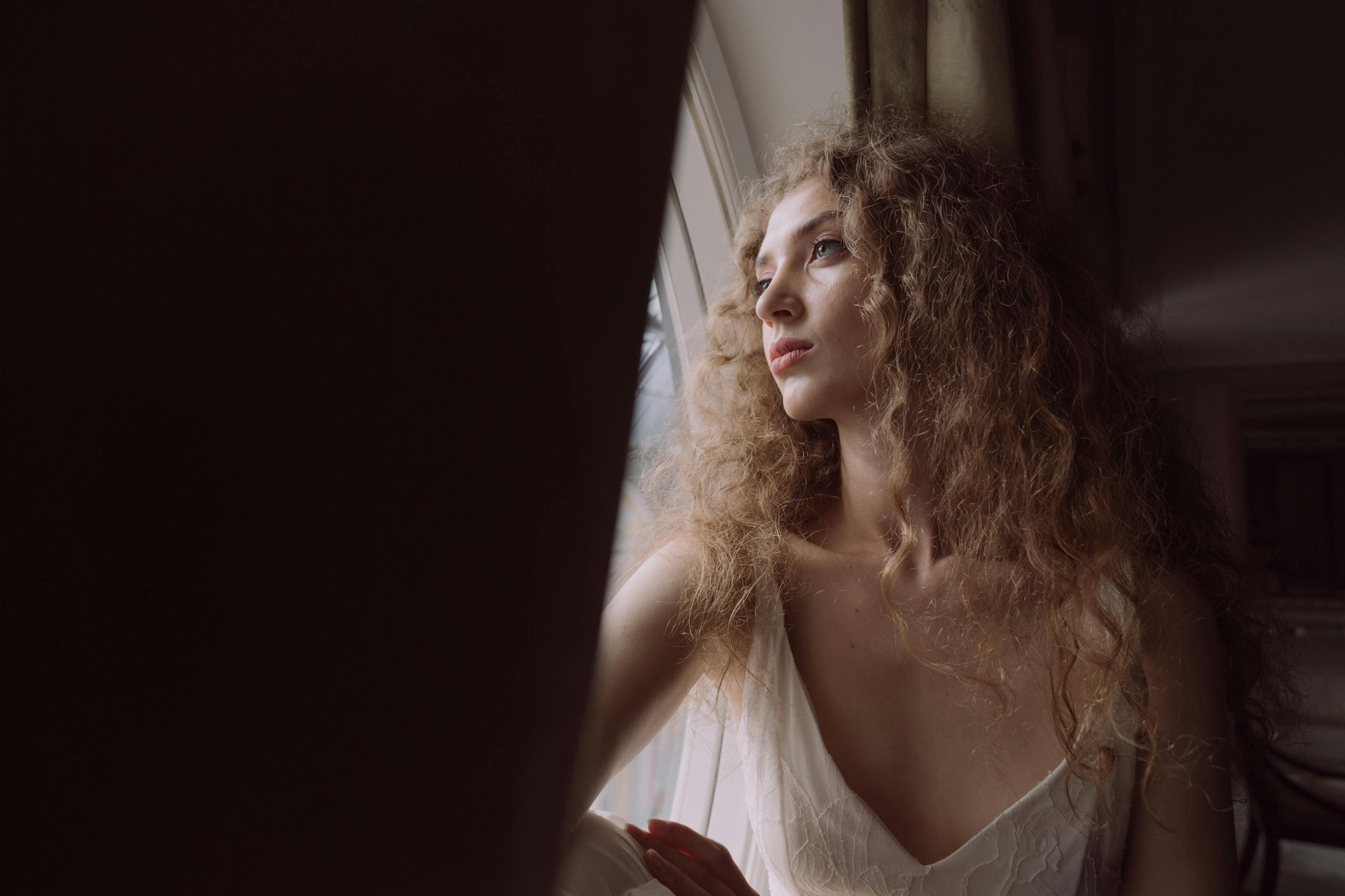 Carmela Clouth A contemplative woman with curly hair looks out a window, creating a serene indoor atmosphere.